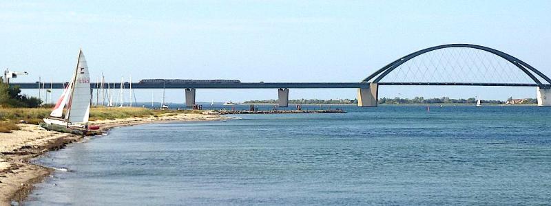 Fehmarnsundbron, vy från Großenbrode, ett tåg passerar bron. 2020 beslutade man att komplettera Fehmarnsundbron med en ny tunnel med motorväg och järnväg. Bron är K-märkt och ska behållas för lokal vägtrafik. I samband med planeringen av Fehmarn Bält-förbindelsen beslutades det att Fehmarnsundbron skulle förbli som den är, med en vägkörbana (och två filer) och ett järnvägsspår, men med elektrifiering tillagd på järnvägen. Foto: Wikipedia Common Lic. Kredit: Holger.Ellgaard -