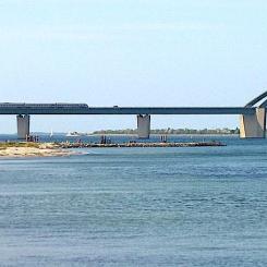 Fehmarnsundbron, vy från Großenbrode, ett tåg passerar bron. 2020 beslutade man att komplettera Fehmarnsundbron med en ny tunnel med motorväg och järnväg. Bron är K-märkt och ska behållas för lokal vägtrafik. I samband med planeringen av Fehmarn Bält-förbindelsen beslutades det att Fehmarnsundbron skulle förbli som den är, med en vägkörbana (och två filer) och ett järnvägsspår, men med elektrifiering tillagd på järnvägen. Foto: Wikipedia Common Lic. Kredit: Holger.Ellgaard -